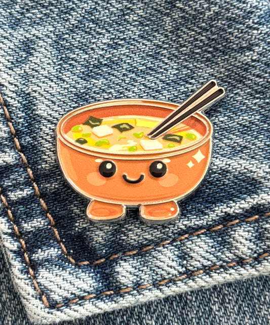A cute patient ramen enamel pin, resting on a blue denim pocket.