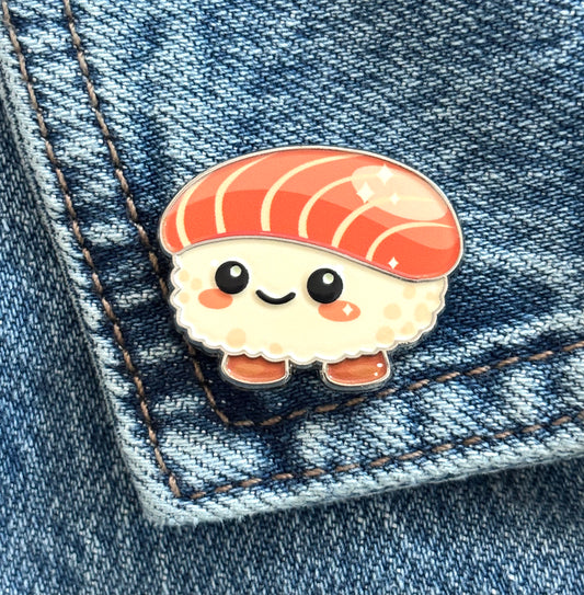 A cute, sweet salmon sushi-shaped enamel pin with a smiling face, resting on a blue denim pocket.