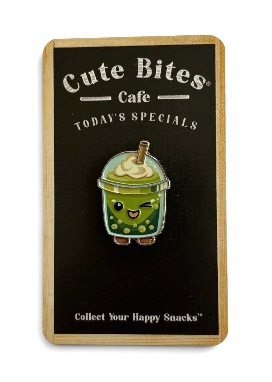 A cute enamel pin of a winking green bubble matcha drink character, mounted on a black display card with a light wood border. The card features the brand name 'Cute Bites Cafe' and the text 'TODAY'S SPECIALS'.