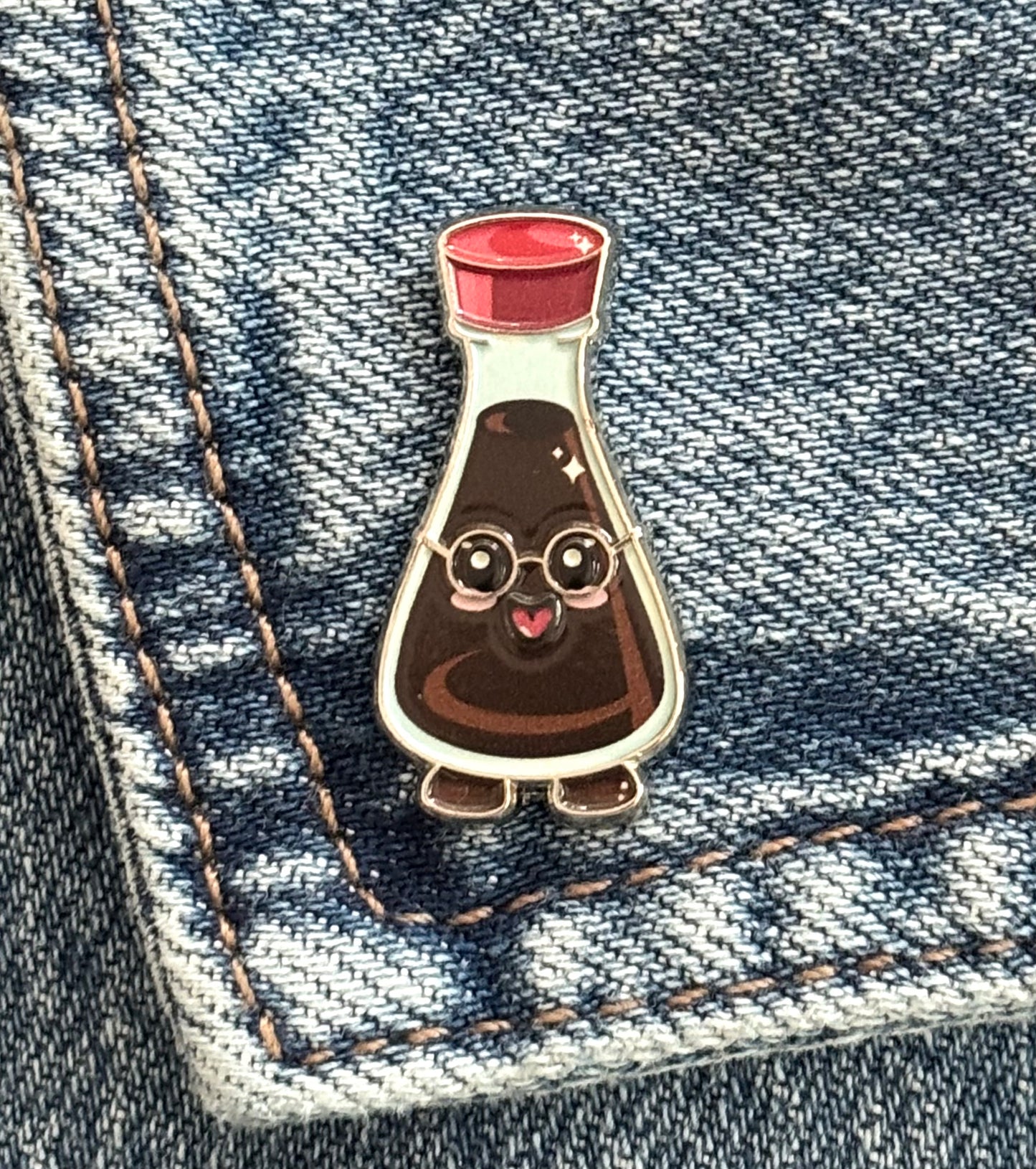 A cute enamel pin of a smart soy bottle with glasses and a red cap, pinned onto a blue denim pocket.