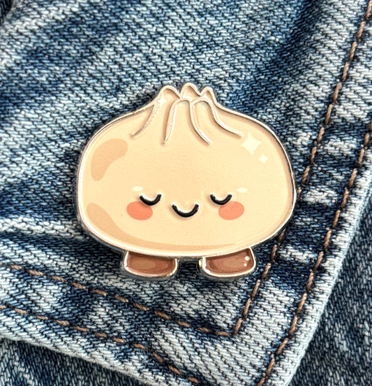 A cute kind dumpling enamel pin with closed eyes and pink cheeks, resting on blue denim fabric.