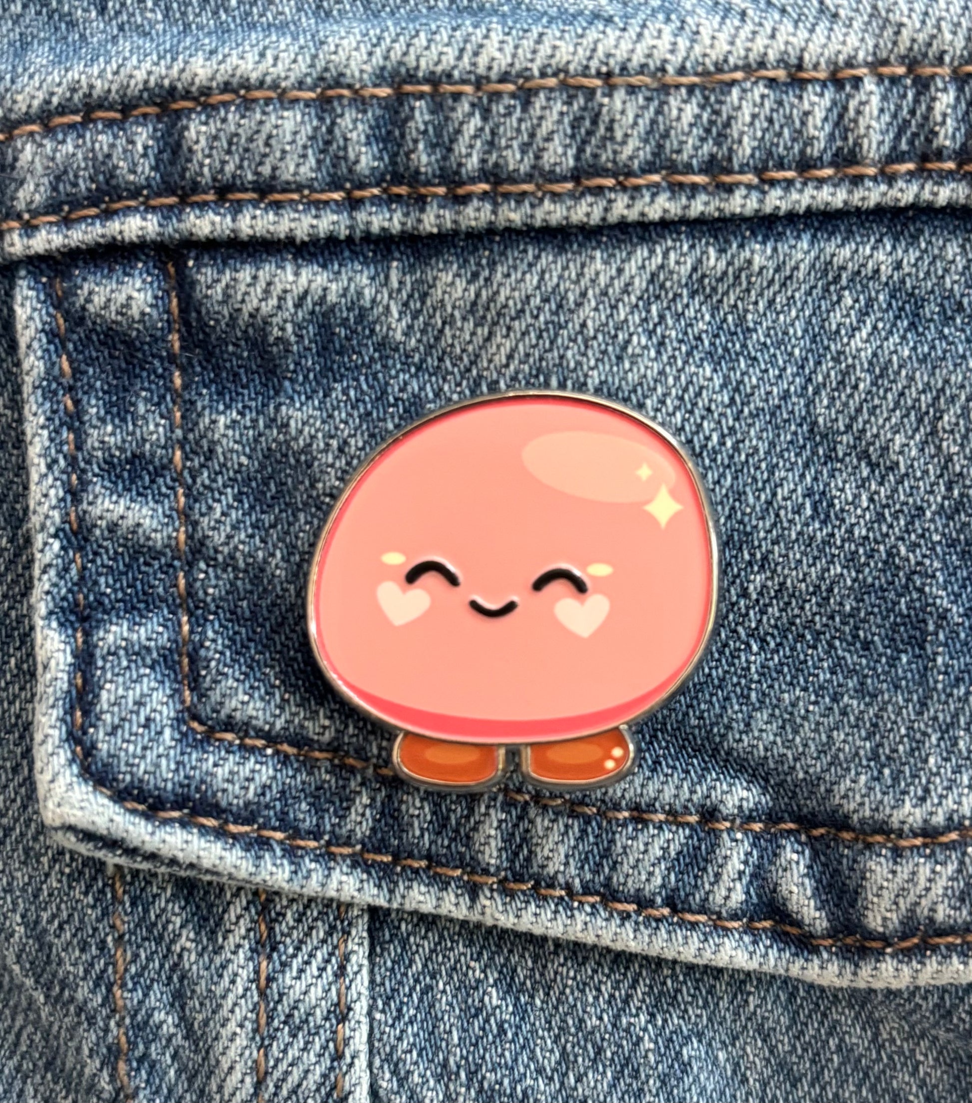 A cute pink gentle mochi enamel pin with a smiling face and small brown feet, pinned onto a blue denim jacket pocket.